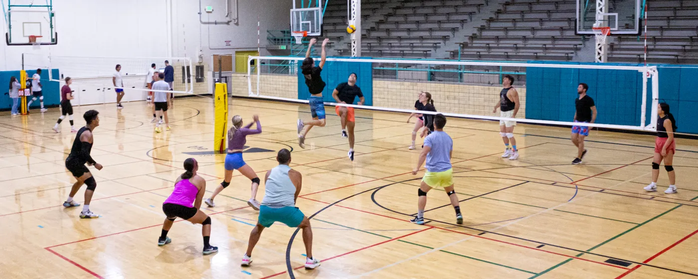 Two volleyball nets are set up side-by-side on an indoor basketball court. Two mixed-gender teams of six young adults face a player leaping high to hit a yellow volleyball just as it sails over the net. The players are dressed for the sport with vibrant athletic clothing and kneepads. Other teams in the background are not actively playing.