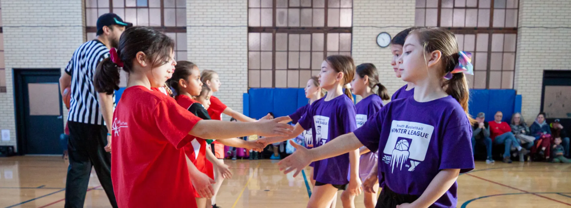 Two teams of five young girl basketball players are lined up in the center of an indoor court. The walls are off-white brick with windows that suggest the building was built in the early 1900s or earlier. The teams are approaching each other with outsteched arms to shake hands at the start of a match as their families sit along the wall to observe the game and a referee in black and white stripes supervises.