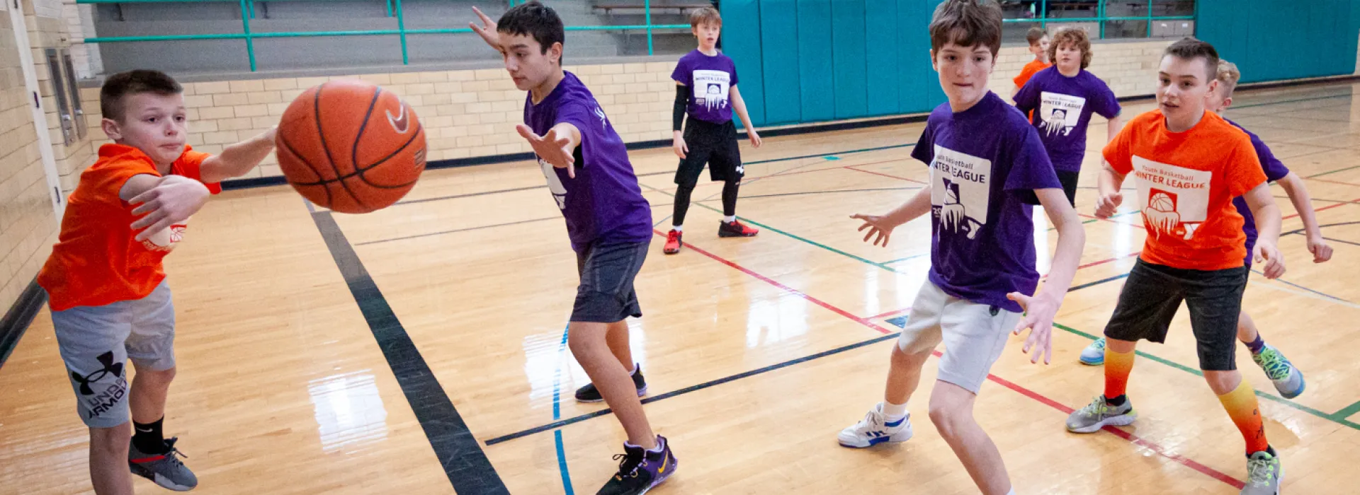 School-aged boys play basketball on an indoor court. Teams are designated with purple or orange shirts. An orange player on the left is tossing the basketball to a player out of frame as the nearest purple player with outstreched hands misses an attempt to block.