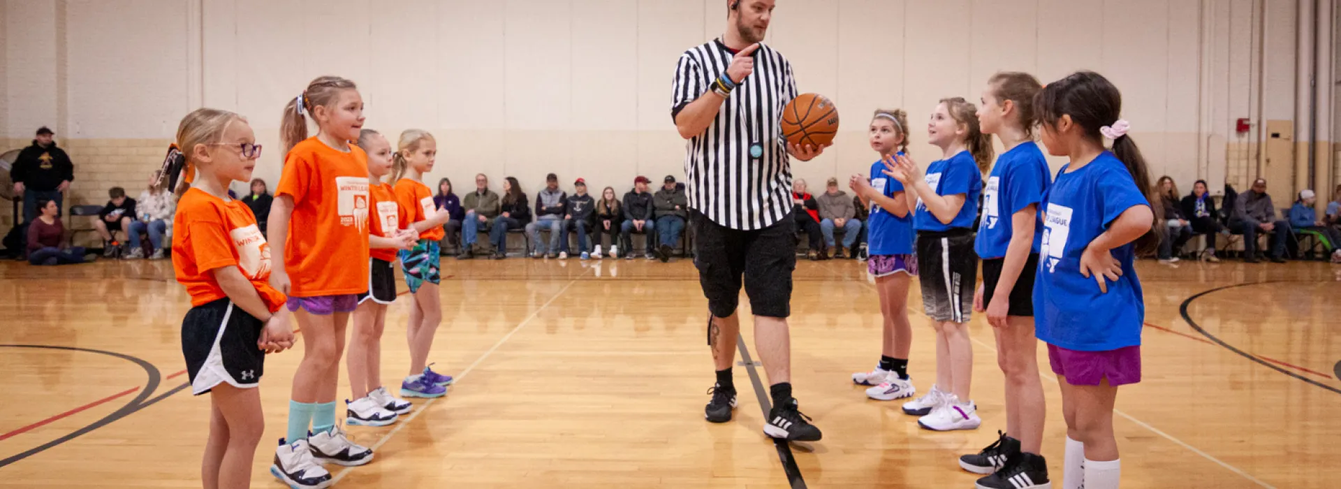 Two teams of four elementary-aged girls line up and face each other at the center of an indoor basketball court. The team on the left wears orange shirts, the team on the left wears blue shirts. Inbetween them walks an adult male referee in a black-and-white striped referee shirt, holding a basketball and speaking. Out of focus along the far wall sits a line of families who are attending the game.